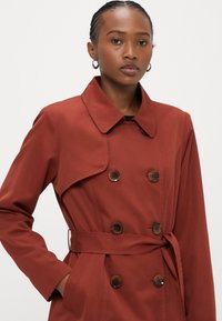 Ženska v rjavo-rjavi jopiči s gumbi v želvjem vzorcu in pasom, stoji z eno roko v žepu na belem ozadju.