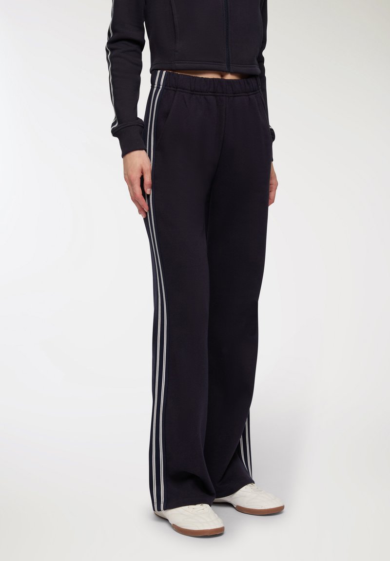 Pantaloni jogger neri realizzati in tessuto morbido, con strisce laterali bianche, vita elastica e tasche laterali. Abbinati a sneaker bianche.