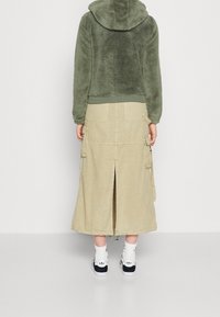 Zielony, puszysty polarowy hoodie z kapturem, zestawiony z długą beżową spódnicą cargo z kieszeniami i rozcięciem z tyłu, noszony z czarnymi sneakerami.