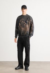 Model iført en sort cap, en mørk sweatshirt med kunstnerisk print af balletdansere, sorte cargo-bukser og sorte blanke sko, står på et gråt gulv.