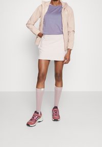 Giacca a vento rosa chiaro sopra una maglietta viola, abbinata a una gonna bianca e calze rosa alte fino al ginocchio, completata con sneakers rosa e bianche.