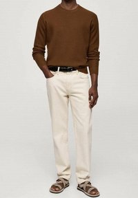 Uomo che indossa un maglione marrone a maniche lunghe, pantaloni color crema con una cintura nera e sandali beige, che si trova in piedi contro uno sfondo neutro.
