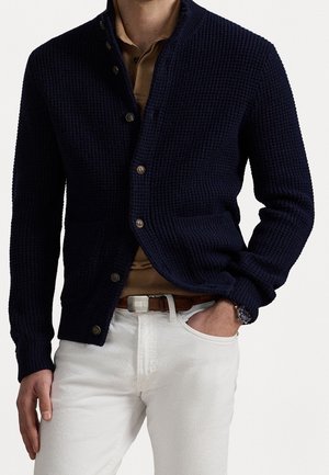 Mann trägt eine marineblaue Strickjacke mit Waffelmuster über einem braunen Hemd, weiße Hose, brauner Gürtel und Uhr, mit einer Hand in der Tasche vor weißem Hintergrund.