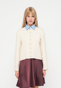 Kremfarget cardigan med en blå skjorte under, med knapper foran og lange ermer, kombinert med et burgunder skjørt.