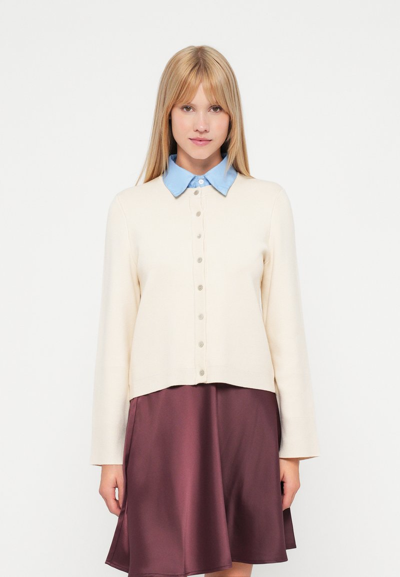 Kremfarget cardigan med en blå skjorte under, med knapper foran og lange ermer, kombinert med et burgunder skjørt.