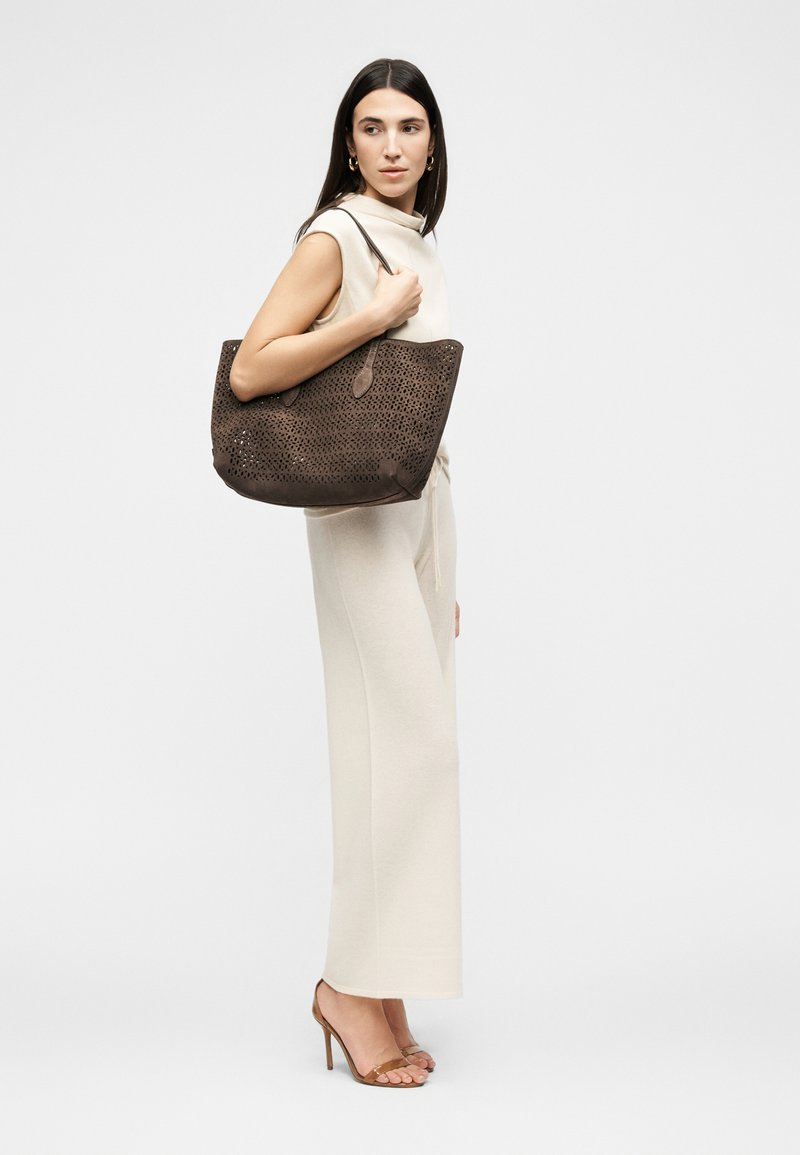 Žena v krémovom tielku bez rukávov a širokých nohaviciach, držiaca veľkú tmavohnedú perforovanú kabelku, nosiaca hnedé sandále na vysokom podpätku, pozerá sa bokom.
