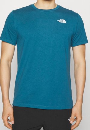 Mężczyzna w turkusowym T-shircie The North Face z okrągłym dekoltem, z logo na górnej lewej części klatki piersiowej, w połączeniu z czarnymi dolnymi częściami z częściowym logo.