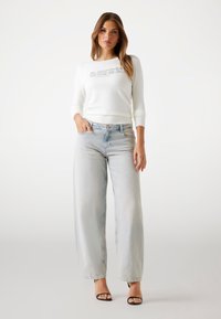 Donna in piedi che indossa una maglietta bianca a maniche lunghe con logo, jeans a gamba larga in denim chiaro e sandali con tacco aperti neri su uno sfondo neutro.