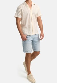 Homme portant une chemise crème à manches courtes boutonnée, un short en jean bleu clair retroussé, et des baskets beige, debout avec une main dans la poche.