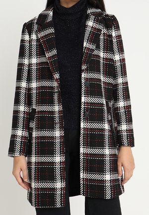 Femme portant un manteau à carreaux noir, blanc et rouge sur un pull à col roulé noir et un pantalon noir.