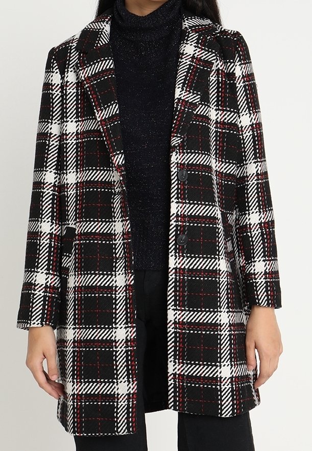 Femme portant un manteau à carreaux noir, blanc et rouge sur un pull à col roulé noir et un pantalon noir.