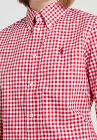 Hedvábná košile v červeno-bílé kostkované vzoru s límečkem na knoflíky, s malým vyšitým logem na hrudi a bílými knoflíky.