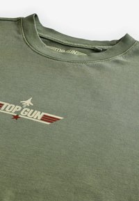 Olivově zelené tričko vyrobené z měkké tkaniny s krémovým logem "TOP GUN" s červenými akcenty a designem letadla pod textem.