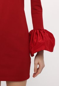 Rochie roșie cu un bust ajustat, cu o textură netedă și mâneci bufante dramatice dintr-un material lucios, creând o siluetă contrastantă.