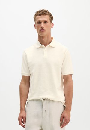Jeune homme portant un polo beige à manches courtes et un pantalon clair à cordon, debout devant un fond blanc uni.