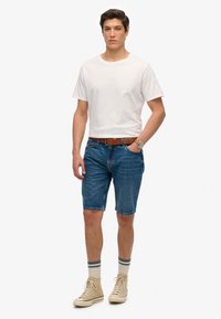 Camiseta blanca de algodón, pantalones cortos de mezclilla azul, cinturón de cuero marrón, zapatillas de caña alta beige con detalles en gris, pose de pie, fondo neutro.