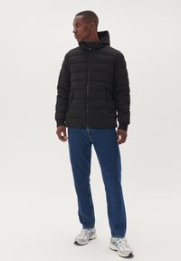 Chaqueta de plumas negra con diseño acolchado horizontal, cierre de cremallera y capucha, combinada con jeans de mezclilla azules y zapatillas grises.