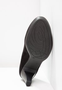 Chaussure en daim noir avec un talon effilé, semelle en caoutchouc texturée présentant un motif en spirale, inclinée contre un fond blanc uni.