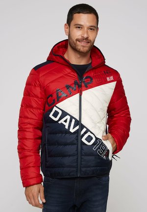 Man wearing a red, white, and navy color-blocked hooded puffer jacket with CAMP DAVID text and zipper pockets, standing against a plain background.