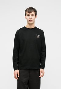 Bărbat tânăr în picioare, îmbrăcat cu un tricou negru cu mânecă lungă, având text în japoneză și o mică petrecere rectangulară pe pieptul stâng.