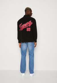 Giacca nera con ricamo rosso "Tommy 85" sulla schiena, abbinata a jeans azzurri e sneaker bianche, in piedi su uno sfondo bianco.
