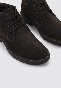 Botas de hombre en ante marrón oscuro hasta el tobillo, con punteras redondas, cierre de cordones de tres ojetes y suelas de goma negra resistentes.