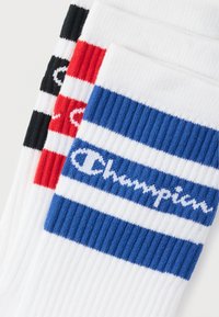 Τρία ζευγάρια λευκές κάλτσες με ρίγες μπλε, κόκκινου και μαύρου χρώματος στην κορυφή και το λογότυπο "Champion" σε λευκό σε κάθε ζευγάρι.