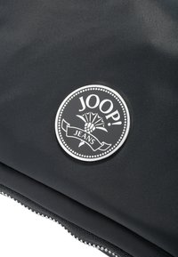 Sacca in tessuto nero con un logo circolare argento recante la scritta "JOOP! JEANS" e dettagli decorativi. Texture liscia, chiusa con una cerniera.