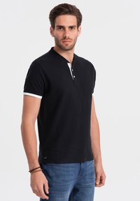 Polo negro con una tapeta de botones blanca y ribetes blancos, hecho de una tela suave. Mangas cortas y corte recto, combinado con unos vaqueros azules.