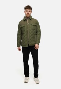 Quiltet olivengrøn jakke med store brystlommer, trykknapper og krave, båret over en beige top og parret med sorte bukser.