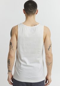 Top din bumbac alb, fără mâneci, cu guler rotund, având un design simplu. Vederea din spate evidențiază tatuajele de pe brațe și gât.