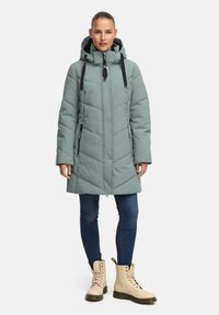 Lange, lichtgroene pufferjas met capuchon, ritssluiting aan de voorkant, zwarte accenten en een gewatteerde textuur. Gecombineerd met blauwe jeans en beige veterschoenen.