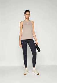 Heledad hallhalli heledast tank top minimalistliku disainiga, tumedate laevaviikidega ja valgete spordijalatsitega; käes must veepudel.