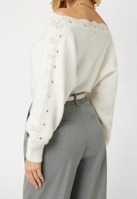 Weißer, gestrickter Pullover mit einem schulterfreien Design, verziert mit Spitzenakzenten an den Ärmeln, kombiniert mit grauen, maßgeschneiderten Hosen und einem beigen Gürtel.