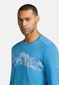 Blå långärmad skjorta i strukturerat tyg, med en vit bergsgrafik över bröstet. Cirkulär halsringning och ribbade detaljer.