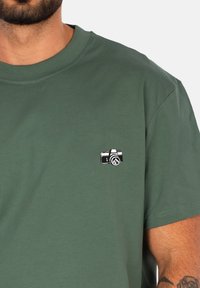 T-shirt in cotone verde con una piccola grafica di una macchina fotografica in bianco e nero sulla parte sinistra del petto. Maniche corte e scollo rotondo.