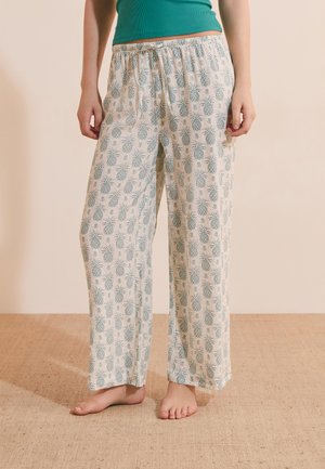 Personne portant un pantalon ample et de couleur claire avec un motif répétitif d'ananas verts, debout pieds nus sur un sol texturé.