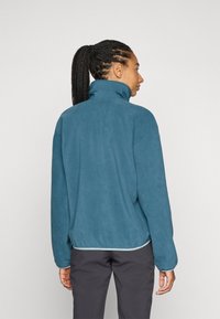 Femme avec des tresses portant une veste en polaire turquoise à col haut, se tenant de dos contre un fond clair et uni.