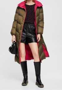 Olivgrüne Pufferjacke mit einer pinken Kapuze, kombiniert mit einem schwarzen Polka-Dot-Top, schwarzen Leder-Shorts und knielangen schwarzen Stiefeln. Schwarze Handtasche.