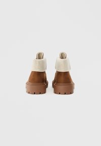 Brune ruskindsankelstøvler med en cremefarvet stofkrave, kraftig brun gummiydersål med synlig profil og et rundt tådesign.