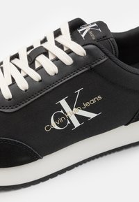 Zapatillas negras con cordones blancos, con una parte superior de textil, una puntera negra suave y un logo dorado de "Calvin Klein Jeans".