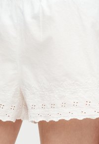 Λευκή φούστα με κεντήματα σε σχήμα λουλουδιών και κυματιστό τελείωμα, φορεμένη από άτομο με ορατά πόδια.