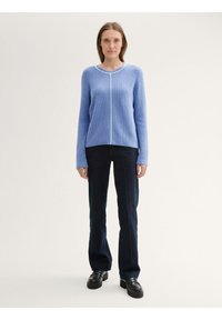 Lichtblauwe gebreide trui met een ronde halslijn, lange mouwen en witte stiknaden langs het midden; gecombineerd met donkerblauwe flared jeans.
