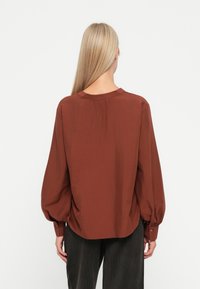 Bruine lange mouwen shirt met een afgeronde zoom, geplooide achterkant en knoopmanchetten. Gemaakt van lichtgewicht stof met een zachte textuur.
