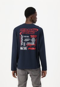 Camiseta de manga larga navy con varios logotipos de Red Bull Racing en rojo y blanco. Material de algodón con un corte holgado y diseño de cuello redondo.