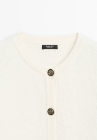 Cardigan bianco lavorato a maglia con scollatura rotonda, caratterizzato da un motivo a rilievo e un bottone dorato con un emblema di leone al centro.