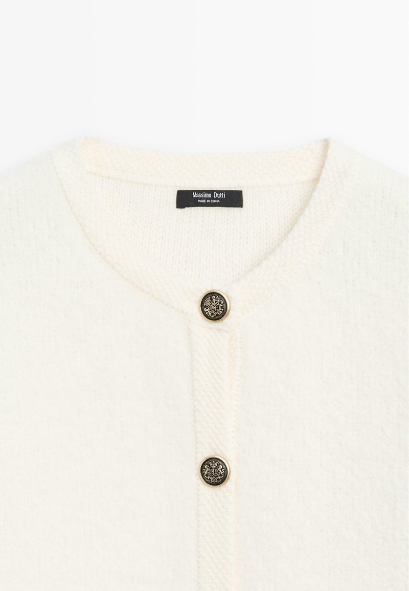 Cardigan bianco lavorato a maglia con scollatura rotonda, caratterizzato da un motivo a rilievo e un bottone dorato con un emblema di leone al centro.