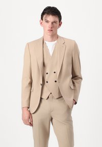Completo beige a tre pezzi realizzato in tessuto liscio; giacca sartoriale, gilet con doppi bottoni e pantaloni aderenti; design minimale.