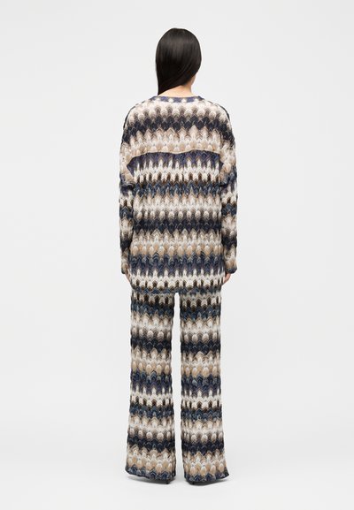 Γυναίκα με μακριά σκούρα μαλλιά, φορόντας ταιριαστό, φαρδύ πλεκτό πουλόβερ και παντελόνια με φαρδιά πόδια σε μπεζ, ναυτικό και λευκό ζιγκ-ζαγκ μοτίβο, εμφανιζόμενη από την πίσω πλευρά.
