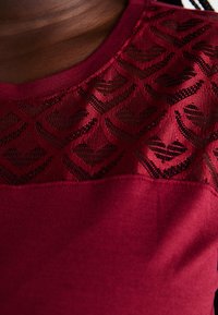 Burgundy top with a textured lace panel featuring a heart pattern. Smooth fabric below the lace with a rounded neckline and short sleeves.